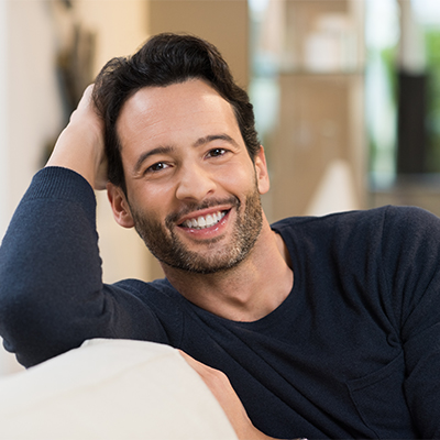 Man with dark hair and beard smiling while sitting on a couch, wearing a dark long-sleeve shirt in a warmly lit indoor setting.