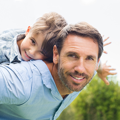 Father giving young son a piggyback ride outdoors, both smiling and looking at the camera in a playful pose.