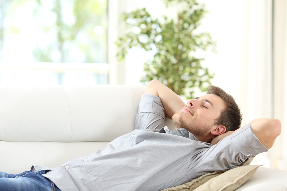 Young man relaxing on a white sofa with eyes closed and hands behind his head, appearing calm and content in a sunlit room.