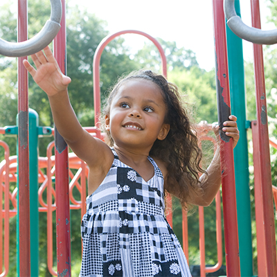 Little girl in a black-and-white patterned dress playing on a playground, holding onto a bar with one hand and reaching out with the other.