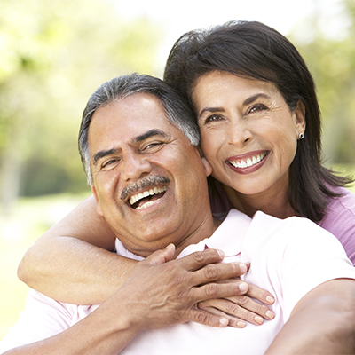 Smiling mature couple embracing outdoors, with the woman hugging the man from behind while both look at the camera.