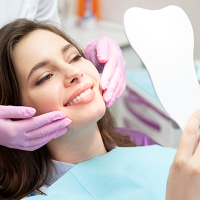 Female dental patient smiling and holding a tooth-shaped mirror while a dental professional wearing pink gloves gently touches her cheeks during a cosmetic dental consultation.