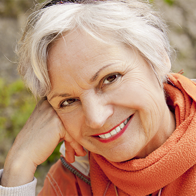 Older woman with short white hair smiling warmly, resting her head on her hand, wearing an orange scarf and sweater outdoors.