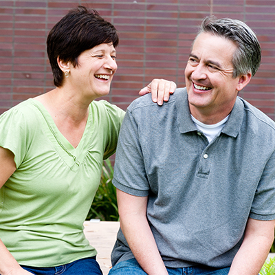Middle-aged couple sitting outdoors and laughing, with the woman in a green top resting her hand on the man's shoulder.
