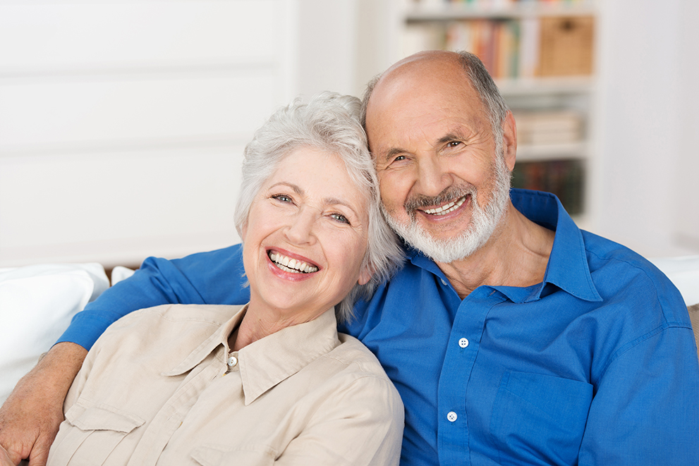 Smiling senior couple sitting close together on a couch, with the woman in a beige shirt and the man in a blue shirt, both looking directly at the camera.