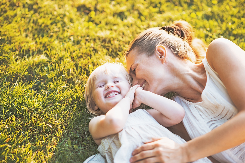 Young mother lying on grass and playfully snuggling her laughing toddler daughter, both dressed in white and bathed in warm sunlight.