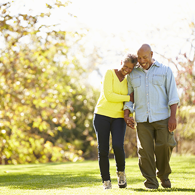Older couple walking arm-in-arm in a sunny park, with the woman in a yellow sweater and the man in a light denim shirt, both smiling and enjoying a relaxed stroll.