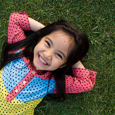 Young girl with long dark hair lying on grass, smiling brightly while resting her hands behind her head and wearing a colorful polka-dot shirt.