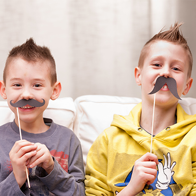 Two young boys sitting on a couch holding fake mustaches on sticks in front of their faces, smiling and playfully posing.