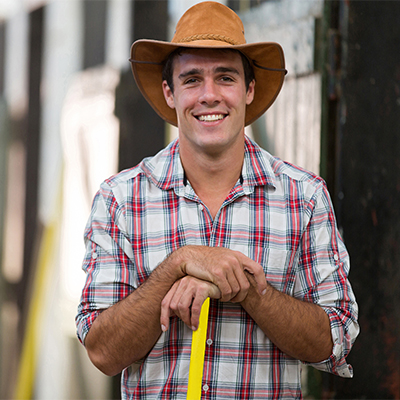 Man in a plaid shirt and brown cowboy hat smiling while leaning on a yellow-handled tool, standing outdoors in front of a barn or stable.