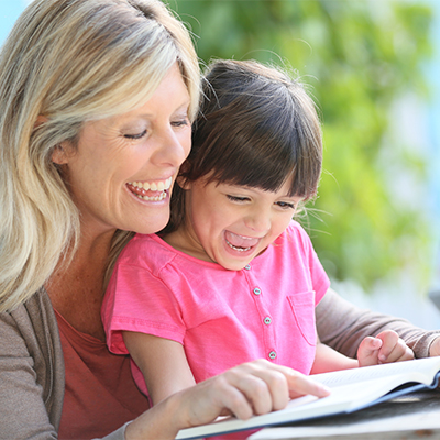 Woman and young girl reading a book together outdoors, both laughing and sharing an engaging moment.