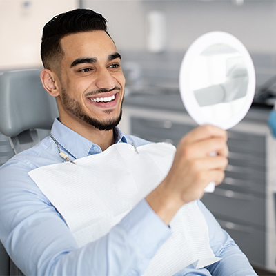 A man seated in a dental chair, holding a mirror and smiling at his own healthy, bright smile.